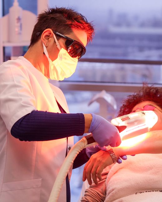 A healthcare professional wearing protective equipment, including a face mask, protective goggles, and gloves, is performing a medical procedure on a patient who is lying on a bed or examination table