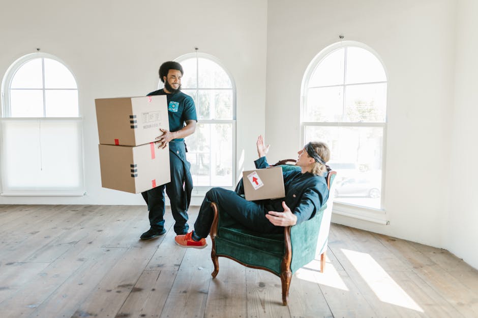 A professional removals worker from Wandsworth Man and Van is inside a bright, spacious room with large, arched windows letting in natural light. The worker, dressed in dark clothing, is carrying two large cardboard boxes stacked on top of each other, secured with red and black packing tape. Nearby, a woman seated on an upholstered vintage armchair with wooden legs, dressed in casual clothing, is holding a third cardboard box with a red arrow sticker on it. She is raising her right hand in a gesture to stop or signal, as if instructing or communicating with the worker, while her left arm rests on the armrest of the chair. The room's wooden floor is clean, and the plain, white walls provide a neutral background. This scene depicts a home relocation or furniture transport process, with packing and moving activities underway. The setting suggests careful handling of items during a house move, illustrating a typical loading and packing stage in house removals by Wandsworth Man and Van.