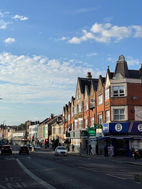A street scene in Wandsworth with a row of multi-story residential buildings featuring brick facades, bay windows, and pitched roofs. In the foreground, there is a busy pavement adjacent to a roadway with several cars and pedestrians. Storefronts and commercial units are visible on the ground level of some buildings, with signs and doors indicating local businesses. Overhead, the sky is partly cloudy with patches of blue, providing natural daylight that illuminates the buildings and street scene. The environment suggests an urban area suitable for house removals or moving services, with potential loading zones on the pavement for furniture transport. Wandsworth Man and Van provides homes and businesses with efficient relocation options contextually aligned with the busy street view.