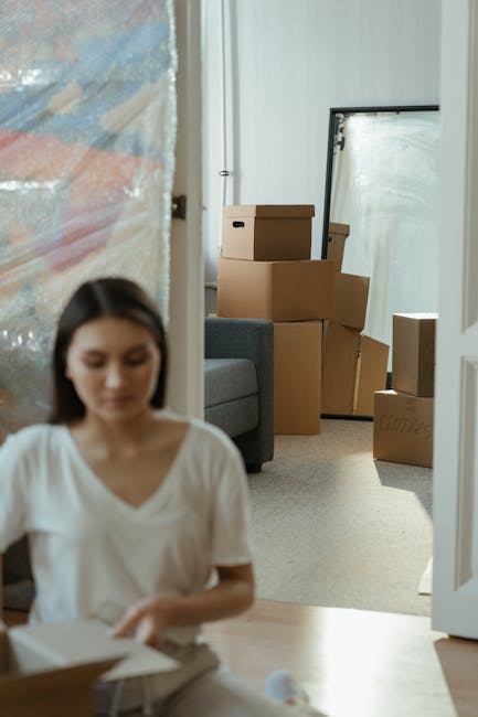 A professional removals worker from Wandsworth Man and Van is inside a bright, spacious room with large, arched windows letting in natural light. The worker, dressed in dark clothing, is carrying two large cardboard boxes stacked on top of each other, secured with red and black packing tape. Nearby, a woman seated on an upholstered vintage armchair with wooden legs, dressed in casual clothing, is holding a third cardboard box with a red arrow sticker on it. She is raising her right hand in a gesture to stop or signal, as if instructing or communicating with the worker, while her left arm rests on the armrest of the chair. The room's wooden floor is clean, and the plain, white walls provide a neutral background. This scene depicts a home relocation or furniture transport process, with packing and moving activities underway. The setting suggests careful handling of items during a house move, illustrating a typical loading and packing stage in house removals by Wandsworth Man and Van.