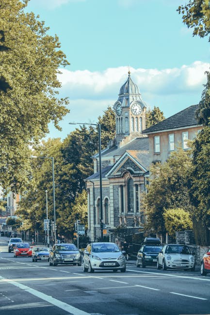 A busy street scene during daytime with multiple parked and moving cars along the roadside, adjacent to a historic building featuring a large clock tower with a pointed spire. The building has ornate architectural details, large arched windows, and a brick exterior. Tall trees with dense foliage line the street, partially shading the sidewalk area. In the background, a crane is visible behind the building, indicating ongoing construction or renovation work. The sky above is clear with some white clouds, and the overall scene captures a typical urban environment suitable for house removals and moving services. Wandsworth Man and Van, a professional removals company, might coordinate such home relocation and furniture transport services within this type of setting.