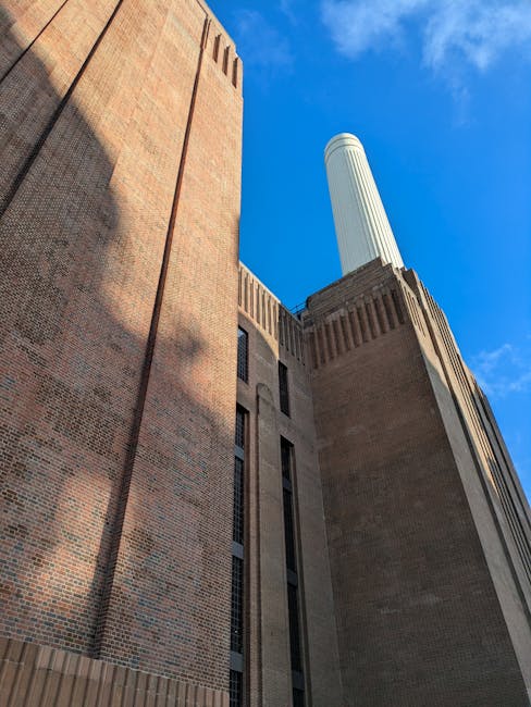 A low-angle view of a large brick building with narrow vertical windows, showing part of a chimney or tower extending upward against a bright blue sky with some wispy clouds. The building appears to be an industrial or institutional structure, with textured brickwork and architectural details near the top. Shadows cast by nearby structures partially shade the brick wall, and the scene captures a clear, sunny day. This image is relevant to house removals and moving services offered by Wandsworth Man and Van, highlighting the context of relocating or transporting items from large or historic buildings as part of a professional home relocation or furniture transport process.